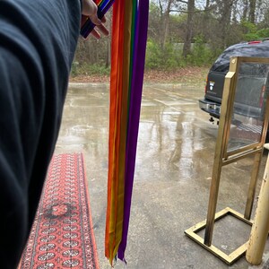 May include: A set of rainbow colored ribbons attached to a set of colorful wands. The ribbons are hanging down and the wands are being held by a hand.