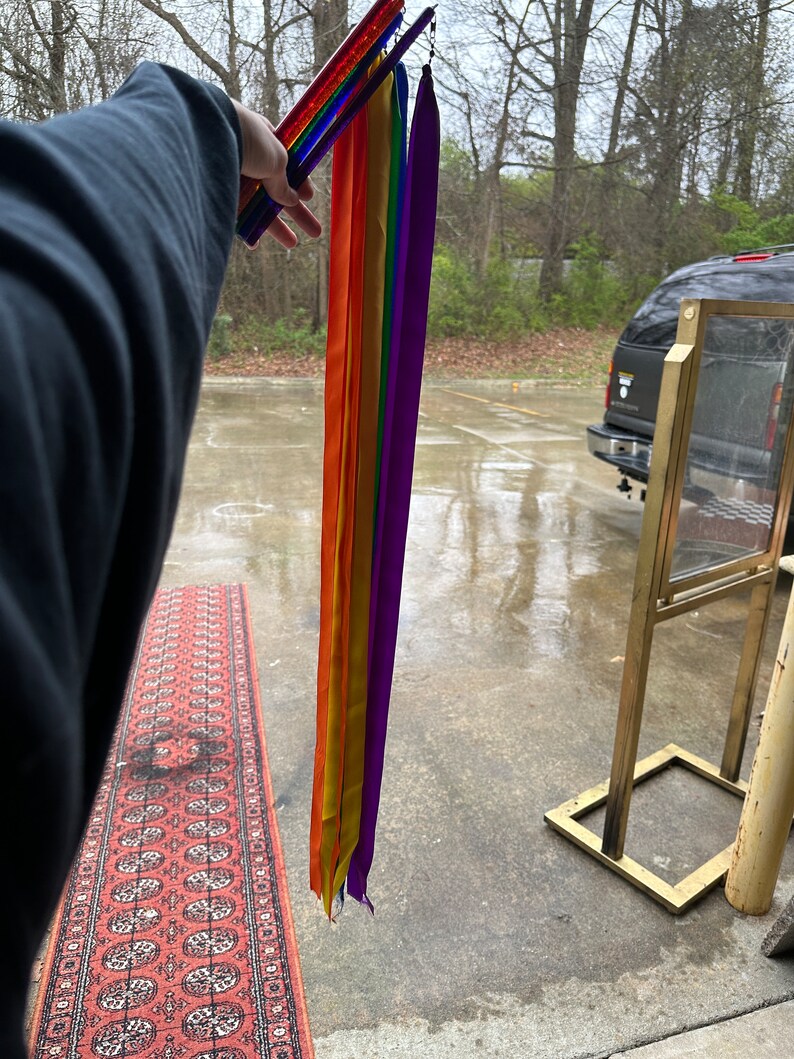 May include: A set of rainbow colored ribbons attached to a set of colorful wands. The ribbons are hanging down and the wands are being held by a hand.