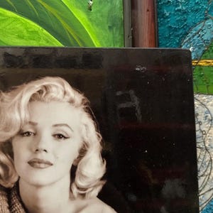 K&ouml;nnte beinhalten: Schwarz-Wei&szlig;-Fotografie von Marilyn Monroe mit ihrem charakteristischen blonden Haar und rotem Lippenstift. Das Bild ist ein Nahportr&auml;t, wobei die Person den Betrachter direkt ansieht. Das Foto ist von einem schwarzen Rahmen umgeben.