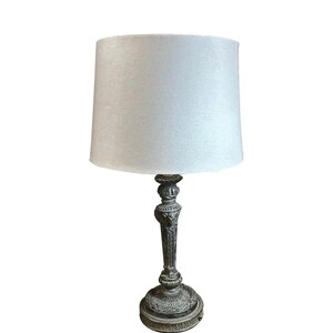 May include: An antique-style table lamp with a white lampshade. The lamp base is ornate, with a detailed, dark metallic finish. The lampshade is a simple, cylindrical shape.