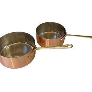 May include: Two copper saucepans with brass handles. The saucepans are nested together, with the smaller saucepan on top.
