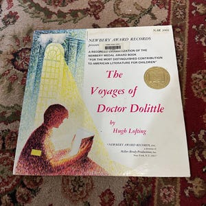 May include: A book cover for "The Voyages of Doctor Dolittle" by Hugh Lofting. The cover features a child reading a book with a window in the background. The book is a recorded dramatization of the Newbery Medal Award book.