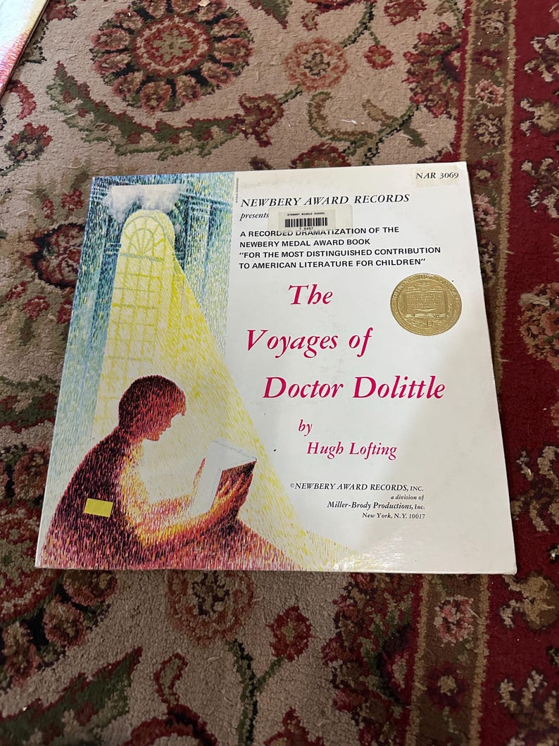 May include: A book cover for "The Voyages of Doctor Dolittle" by Hugh Lofting. The cover features a child reading a book with a window in the background. The book is a recorded dramatization of the Newbery Medal Award book.