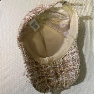 May include: A tweed baseball cap in shades of cream, gold, and brown. The cap has a structured brim and a soft, cream-colored interior lining. The fabric has a textured, woven appearance with metallic threads.