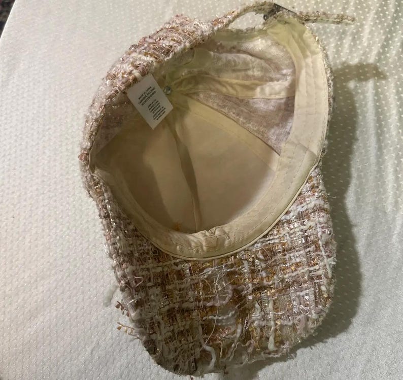 May include: A tweed baseball cap in shades of cream, gold, and brown. The cap has a structured brim and a soft, cream-colored interior lining. The fabric has a textured, woven appearance with metallic threads.