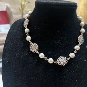 Puede incluir: Un collar dorado con perlas de imitaci&oacute;n y adornos de filigrana plateada. El collar se exhibe en un maniqu&iacute; negro, mostrando su dise&ntilde;o y detalles. Es una pieza de joyer&iacute;a.