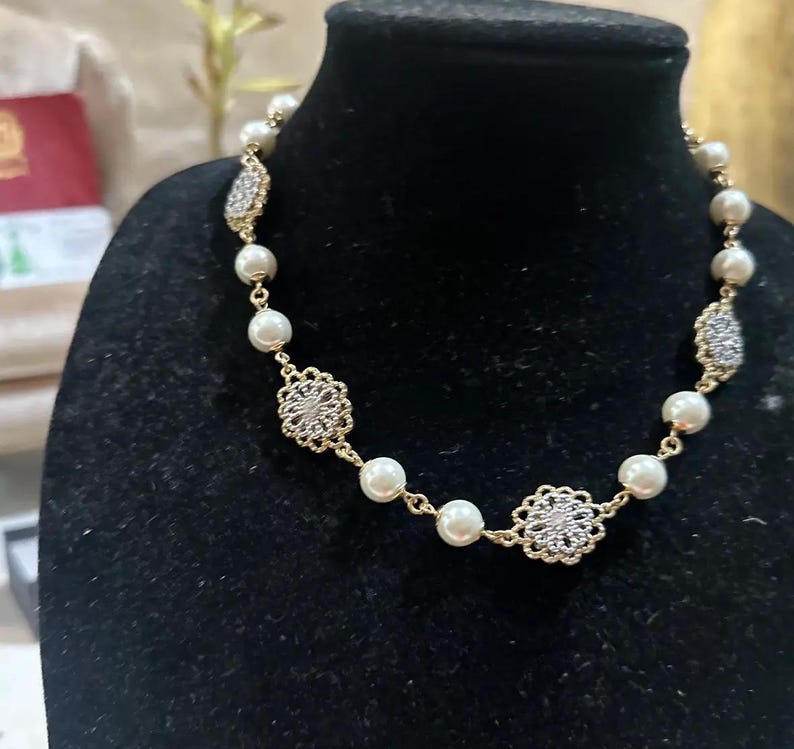 Puede incluir: Un collar dorado con perlas de imitaci&oacute;n y adornos de filigrana plateada. El collar se exhibe en un maniqu&iacute; negro, mostrando su dise&ntilde;o y detalles. Es una pieza de joyer&iacute;a.