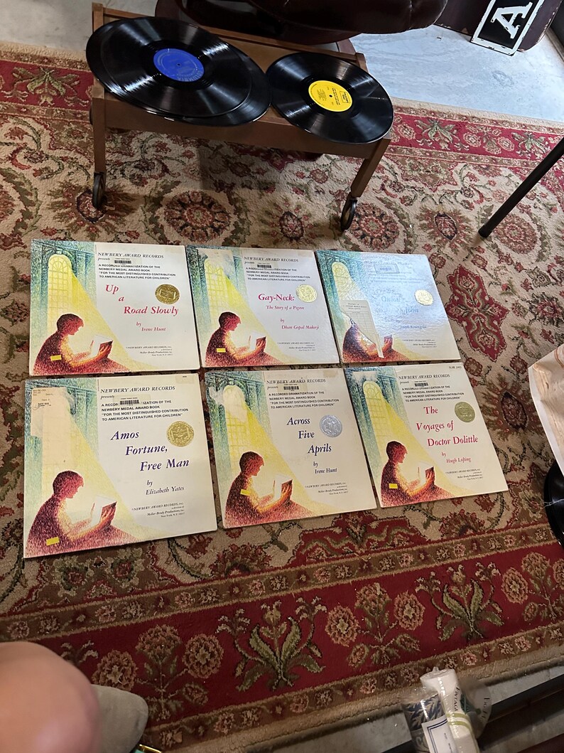 May include: Six vintage Newbery Award Records featuring illustrations of children reading books. The titles are "Up a Road Slowly", "Gay-Neck: The Tale of a Pigeon", "Amos Fortune, Free Man", "Across Five Aprils", "The Voyages of Doctor Dolittle", and "The Story of a Bad Boy".