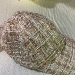 May include: A close-up of a baseball cap made of a woven fabric with a mix of white, pink, and gold threads. The cap has a textured appearance with a slight sheen from the interwoven threads.
