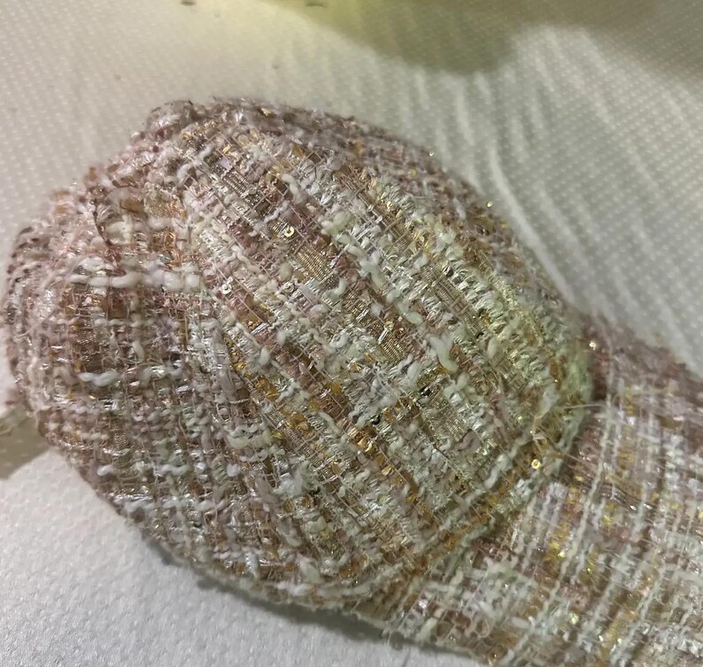 May include: A close-up of a baseball cap made of a woven fabric with a mix of white, pink, and gold threads. The cap has a textured appearance with a slight sheen from the interwoven threads.