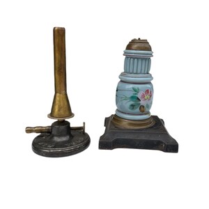 Puede incluir: Dos lámparas antiguas de estilo quemador Bunsen de latón y hierro fundido. Una lámpara tiene una base de porcelana azul y blanca con un diseño floral. La otra lámpara tiene una base negra con un quemador de latón.