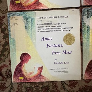 May include: A vintage record album cover featuring a child reading a book. The cover is a light pink and yellow color with a gold seal. The title of the album is "Amos Fortune, Free Man" by Elizabeth Yates. The album is a Newbery Award Record.