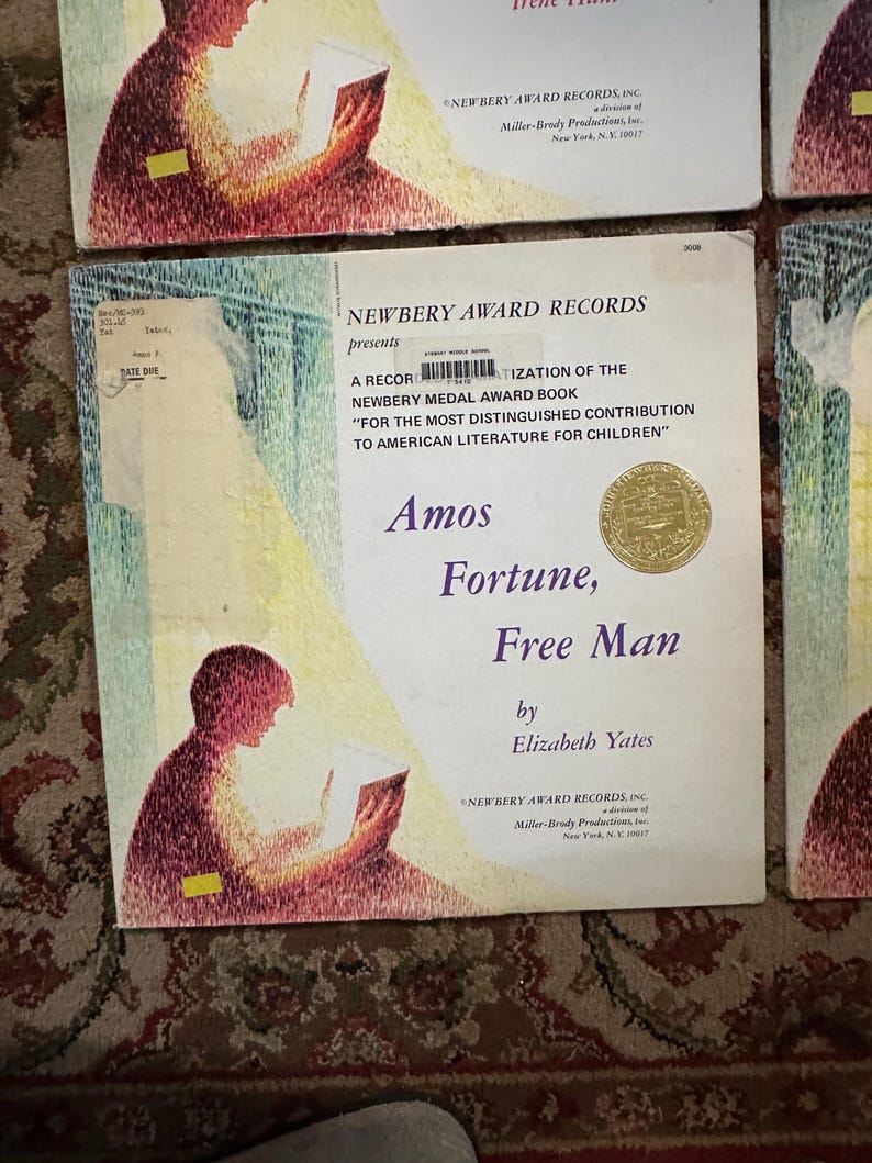 May include: A vintage record album cover featuring a child reading a book. The cover is a light pink and yellow color with a gold seal. The title of the album is "Amos Fortune, Free Man" by Elizabeth Yates. The album is a Newbery Award Record.