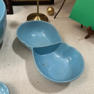 May include: A light blue ceramic divided serving dish. The dish has two connected oval-shaped compartments. The dish is on a light-colored surface. The dish is likely vintage or retro.