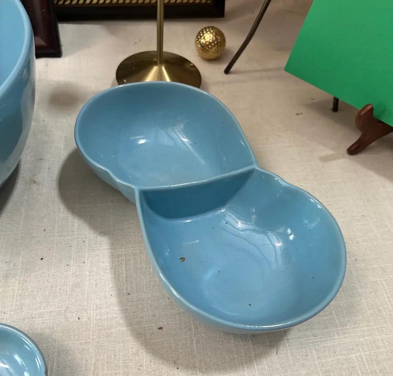 May include: A light blue ceramic divided serving dish. The dish has two connected oval-shaped compartments. The dish is on a light-colored surface. The dish is likely vintage or retro.