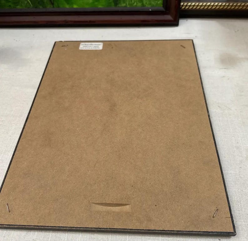 K&ouml;nnte beinhalten: R&uuml;ckseite eines gerahmten Bildes mit brauner Kartonr&uuml;ckwand. Der Rahmen ist dunkelbraun mit einem schwarzen Rand. Ein kleiner wei&szlig;er Aufkleber ist oben mittig auf dem Karton angebracht. Ein horizontaler Schlitz ist unten mittig in den Karton geschnitten.