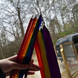 May include: A hand holds a set of rainbow-colored glitter wands with attached rainbow-colored streamers. The wands are made of clear plastic and have a sparkly finish.