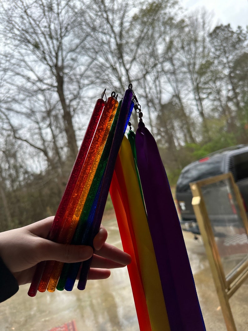 May include: A hand holds a set of rainbow-colored glitter wands with attached rainbow-colored streamers. The wands are made of clear plastic and have a sparkly finish.