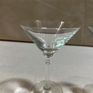 May include: A clear martini glass with a triangular bowl and a round base. The glass is filled with a clear liquid, reflecting light. The stem is slender and elegant, and the glass is set against a neutral background.