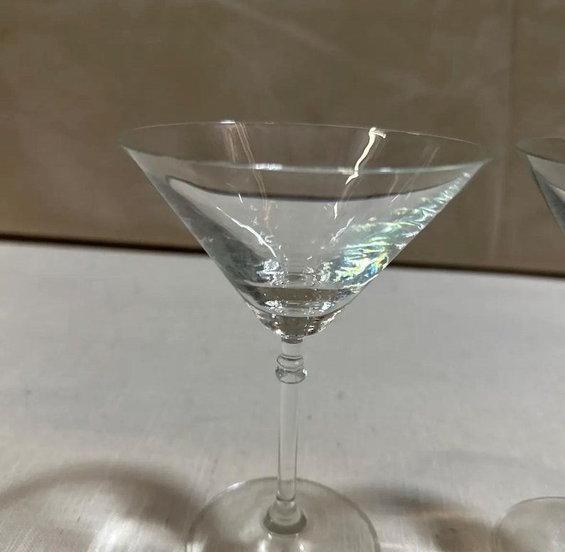 May include: A clear martini glass with a triangular bowl and a round base. The glass is filled with a clear liquid, reflecting light. The stem is slender and elegant, and the glass is set against a neutral background.