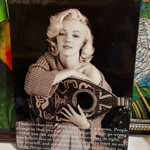 K&ouml;nnte beinhalten: Schwarz-Wei&szlig;-Druck mit Marilyn Monroe, die ein Saiteninstrument h&auml;lt. Das Bild enth&auml;lt ein Zitat von Monroe: "Ich glaube, dass alles aus einem bestimmten Grund geschieht..." Der Druck hat einen dunklen Hintergrund und ist gerahmt.
