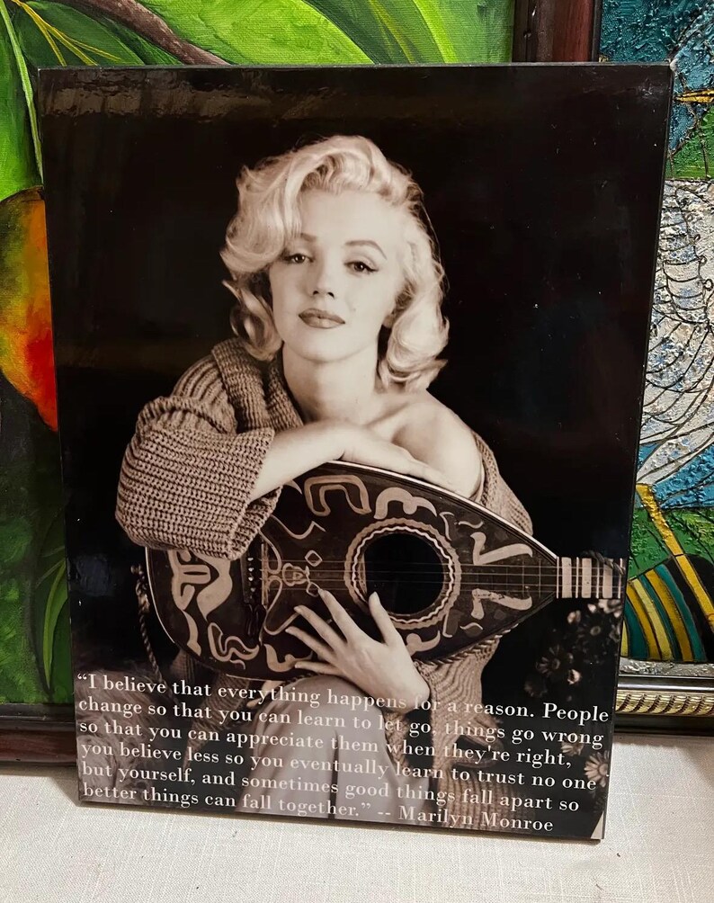 K&ouml;nnte beinhalten: Schwarz-Wei&szlig;-Druck mit Marilyn Monroe, die ein Saiteninstrument h&auml;lt. Das Bild enth&auml;lt ein Zitat von Monroe: "Ich glaube, dass alles aus einem bestimmten Grund geschieht..." Der Druck hat einen dunklen Hintergrund und ist gerahmt.
