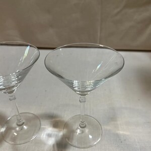 May include: Two clear martini glasses with a classic conical bowl and a slender stem. The glasses are made of transparent glass, reflecting light. The stem connects to a round base. Ideal for cocktails.