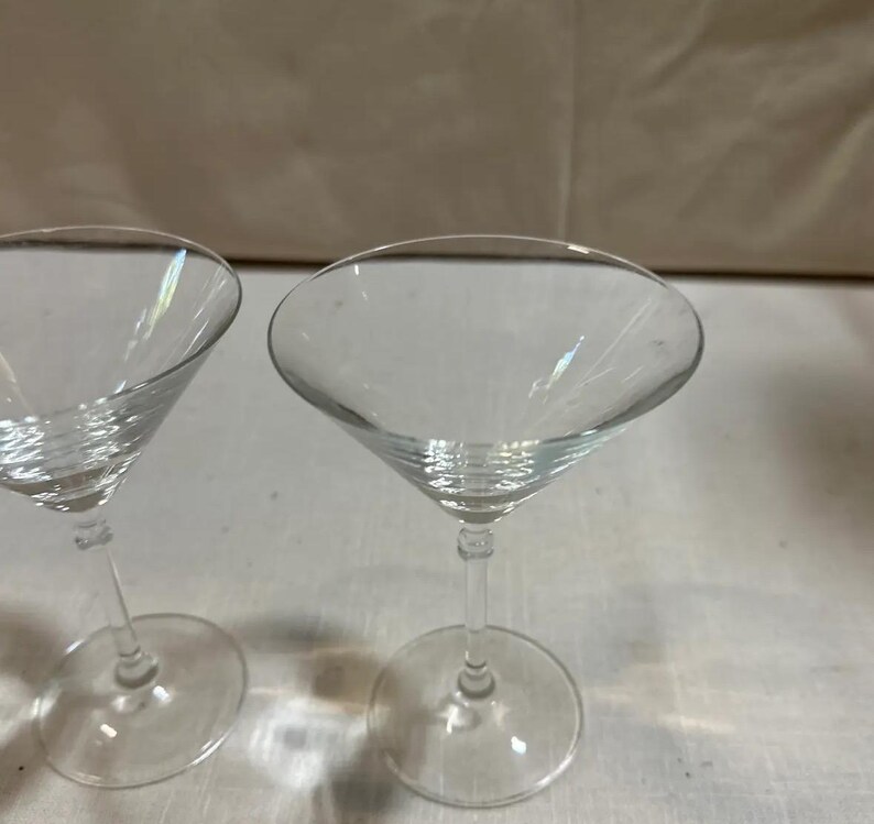May include: Two clear martini glasses with a classic conical bowl and a slender stem. The glasses are made of transparent glass, reflecting light. The stem connects to a round base. Ideal for cocktails.
