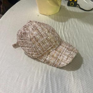 May include: A beige tweed baseball cap with a curved brim. The cap has a textured appearance with subtle sequin accents. The hat is made of a woven fabric and is designed for casual wear.
