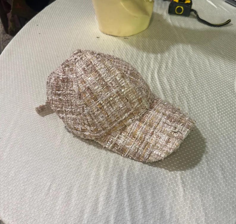 May include: A beige tweed baseball cap with a curved brim. The cap has a textured appearance with subtle sequin accents. The hat is made of a woven fabric and is designed for casual wear.