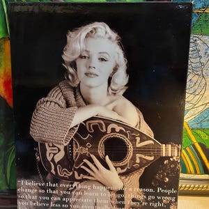 K&ouml;nnte beinhalten: Schwarz-Wei&szlig;-Druck mit Marilyn Monroe, die eine Laute h&auml;lt. Das Bild enth&auml;lt ein Zitat von Monroe: "Ich glaube, dass alles aus einem Grund geschieht..." Der Druck hat einen dunklen Hintergrund und ist gerahmt.
