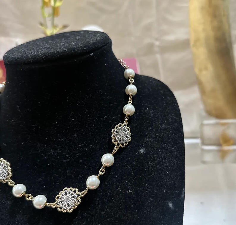 Puede incluir: Un collar dorado con perlas de imitaci&oacute;n y colgantes florales calados con cristales incrustados. El collar se exhibe sobre un busto de terciopelo negro, mostrando su delicado dise&ntilde;o y elegante estilo.