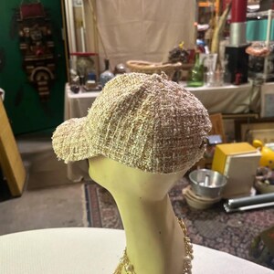 May include: A beige tweed baseball cap with a curved brim. The cap is displayed on a mannequin head with a gold necklace. The cap has a woven texture and a classic baseball cap design.