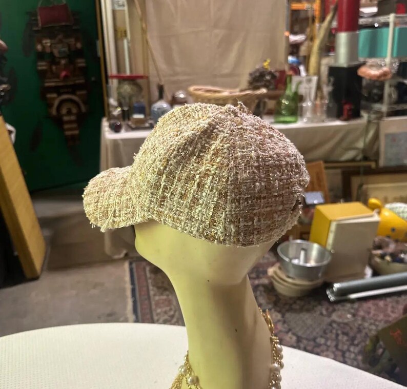 May include: A beige tweed baseball cap with a curved brim. The cap is displayed on a mannequin head with a gold necklace. The cap has a woven texture and a classic baseball cap design.