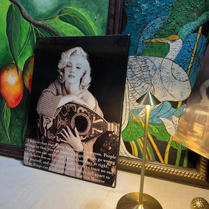 K&ouml;nnte beinhalten: Schwarz-Wei&szlig;-Druck von Marilyn Monroe mit Mandoline und Zitat. Das Bild ist vor einem schwarzen Hintergrund zu sehen. Im Hintergrund sind weitere gerahmte Kunstwerke sowie eine goldfarbene Lampe zu sehen.