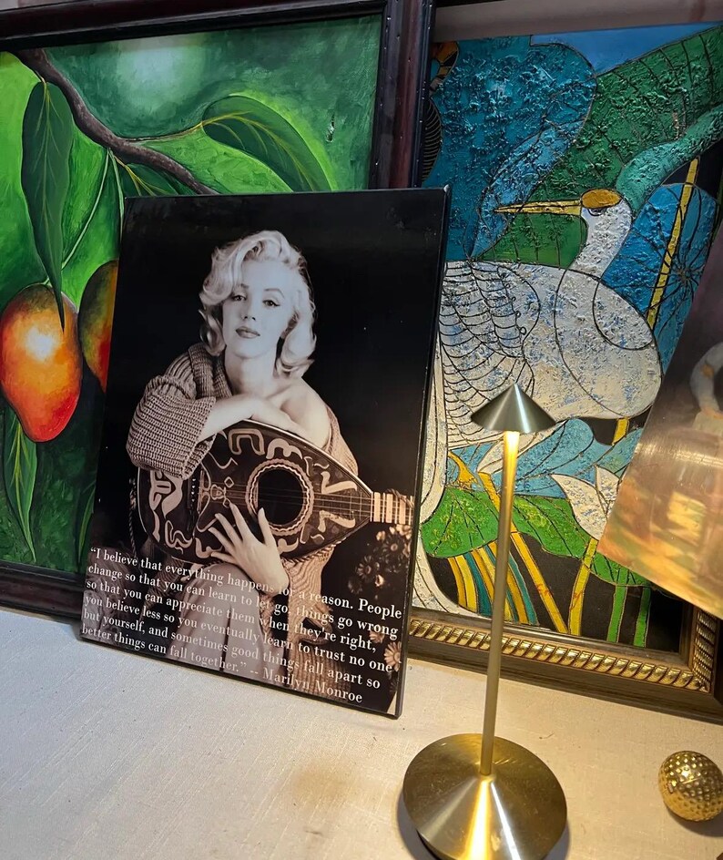 K&ouml;nnte beinhalten: Schwarz-Wei&szlig;-Druck von Marilyn Monroe mit Mandoline und Zitat. Das Bild ist vor einem schwarzen Hintergrund zu sehen. Im Hintergrund sind weitere gerahmte Kunstwerke sowie eine goldfarbene Lampe zu sehen.