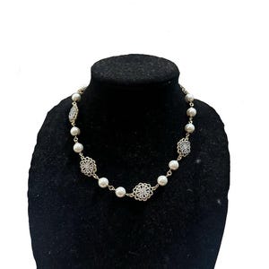 Puede incluir: Un collar de perlas con detalles dorados y elementos decorativos de filigrana. El collar presenta perlas blancas redondas, intercaladas con dise&ntilde;os met&aacute;licos calados y ornamentados. El collar se exhibe sobre un maniqu&iacute; negro.