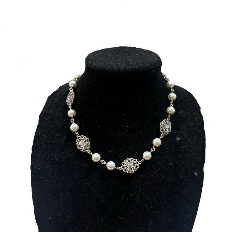 Puede incluir: Un collar de perlas con detalles dorados y elementos decorativos de filigrana. El collar presenta perlas blancas redondas, intercaladas con dise&ntilde;os met&aacute;licos calados y ornamentados. El collar se exhibe sobre un maniqu&iacute; negro.
