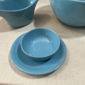 May include: A collection of light blue ceramic dishware, including a small bowl on a matching plate, a gravy boat, and a large bowl. The pieces have a smooth, glossy finish and a mid-century modern design.