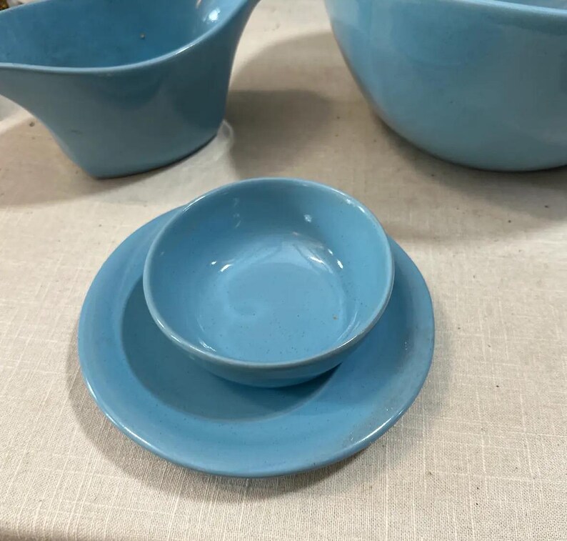 May include: A collection of light blue ceramic dishware, including a small bowl on a matching plate, a gravy boat, and a large bowl. The pieces have a smooth, glossy finish and a mid-century modern design.