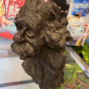 May include: A dark brown ceramic sculpture of a man's head with a textured surface. The sculpture has a large nose and a detailed beard.