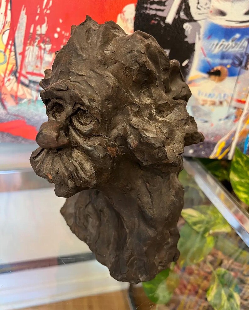 May include: A dark brown ceramic sculpture of a man's head with a textured surface. The sculpture has a large nose and a detailed beard.