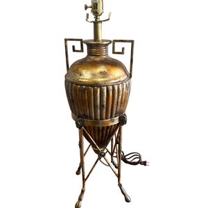 May include: A decorative table lamp with a gold and bronze finish. The lamp features a vase-shaped body with vertical grooves, supported by a tripod base. The lamp has a brass-colored socket and a power cord.