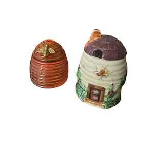 Könnte beinhalten: Zwei Keramik-Honigtopfbehälter. Einer ist in Form eines Bienenstocks mit einer goldenen Biene auf der Oberseite. Der andere ist in Form eines Hauses mit einem violetten Dach und einer Biene an der Seite.