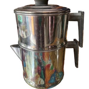 Op de afbeelding: Een roestvrijstalen percolator met een cilindrisch lichaam, een puntige tuit en twee gestapelde grijze handvatten aan de zijkant. Het deksel is voorzien van een donkergrijze knop. Het gepolijste metalen oppervlak reflecteert kleurrijke omgevingen.