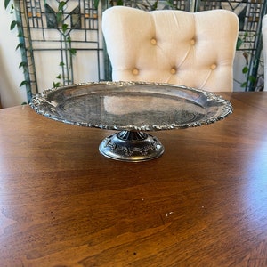 May include: A silver cake stand with a decorative edge and a pedestal base. The stand is on a wooden table.