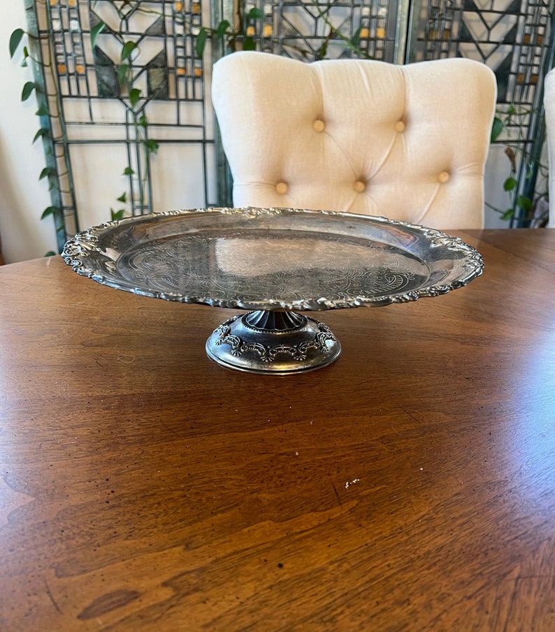 May include: A silver cake stand with a decorative edge and a pedestal base. The stand is on a wooden table.