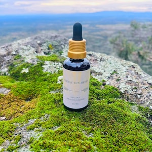 Op de afbeelding: Een kleine, donkere glazen fles met een zwarte druppelaar en goudkleurige dop, met het label "Forest Sun Herbs Organic Dragons Blood". De fles staat op een met mos bedekte rots met een vage achtergrond. Bevat 30 ml vloeistof.