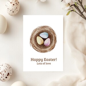 Op de afbeelding: Paaskaart met een aquarel illustratie van een nest met drie pastelkleurige eieren. De kaart heeft de tekst "Happy Easter! Lots of love". Witte en gespikkelde eieren liggen verspreid.