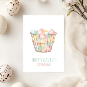 Op de afbeelding: Een paas wenskaart met een draadmand vol pastelkleurige eieren. De kaart toont de tekst "HAPPY EASTER! LOTS OF LOVE" in lichtblauwe letters. Witte en gespikkelde eieren zijn rondom de kaart geplaatst.